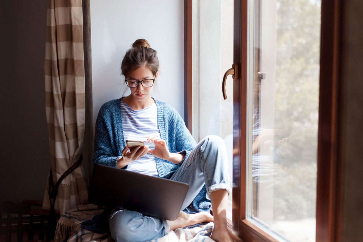 A young woman wearing glasses sits by a window looking at a cell phone in her hands. A laptop rests on her leg.