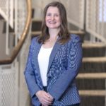 A woman with long hair and wearing a skirt, shirt and blazer smiles.