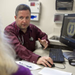 A man in a dress shirt with a stethoscope around his neck sits at a computer in a clinic room. He talks with a woman who is in the foreground, facing away from the camera. A lung scan is on the computer screen.