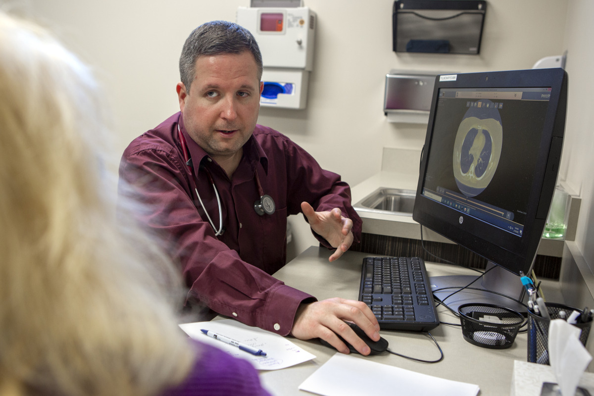 A man in a dress shirt with a stethoscope around his neck sits at a computer in a clinic room. He talks with a woman who is in the foreground, facing away from the camera. A lung scan is on the computer screen.