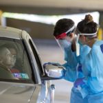 Two people in protective equipment stand at the window of a car in a parking garage.