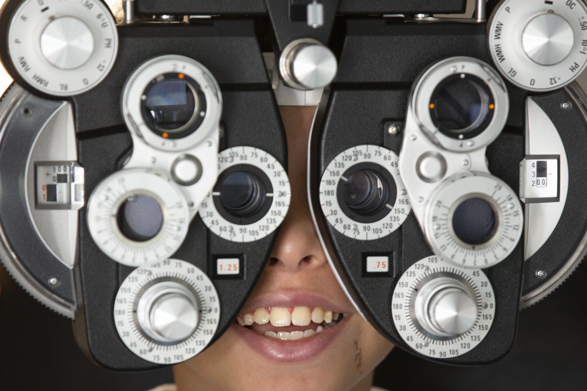 A close-up of a young child looking through the lenses of eye examination equipment, which contains several dials.