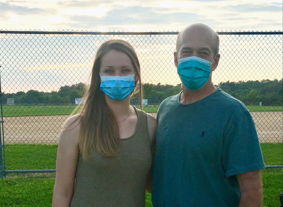 Shalisa Walker stands arm in arm with John Dohner in front of a chain link fence at a softball field. Walker has long hair and wears a tank top. Dohner, who is bald, wears a T-shirt. Both are wearing masks.