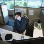 Angie Sellers wears a surgical mask and sits at a computer in a room full of empty cubicles. Behind her, through a window, trees line a parking lot.