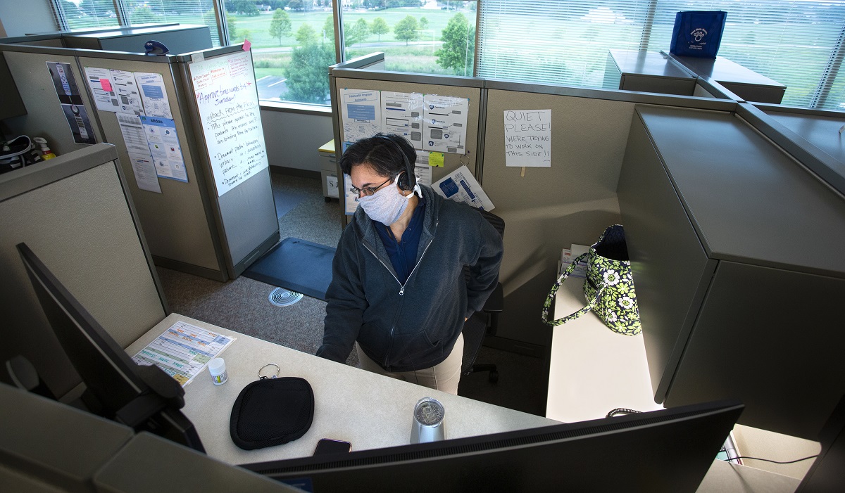 Angie Sellers wears a surgical mask and sits at a computer in a room full of empty cubicles. Behind her, through a window, trees line a parking lot.