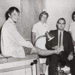 Dr. Willis W. Willard III, wearing a suit and tie, holds the foot of a young boy on an exam table. Two nurses, dressed in white dresses and nursing caps, stand in the background. The boy wears shorts and a jacket and is smiling.