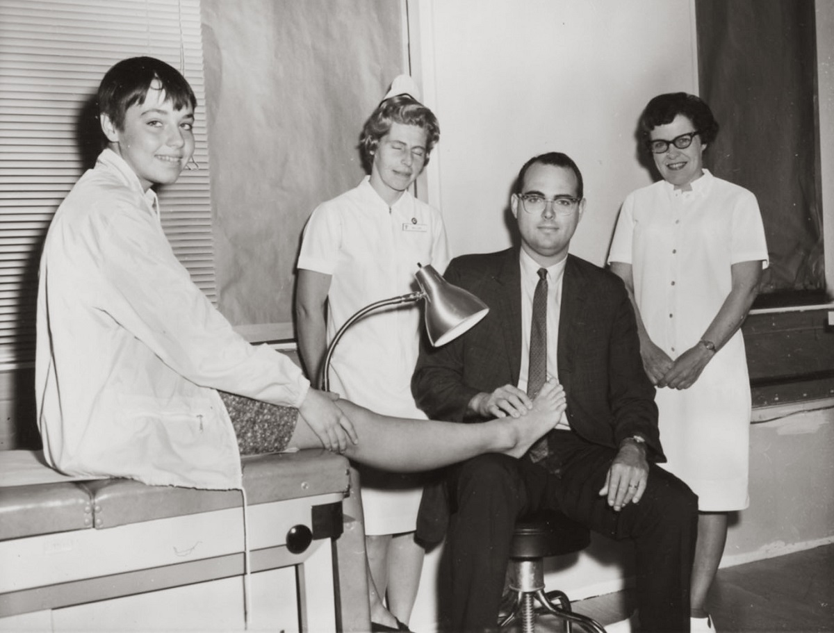 Dr. Willis W. Willard III, wearing a suit and tie, holds the foot of a young boy on an exam table. Two nurses, dressed in white dresses and nursing caps, stand in the background. The boy wears shorts and a jacket and is smiling.
