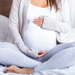 A pregnant woman sits cross-legged, facing the camera, her hands placed above and below her stomach.