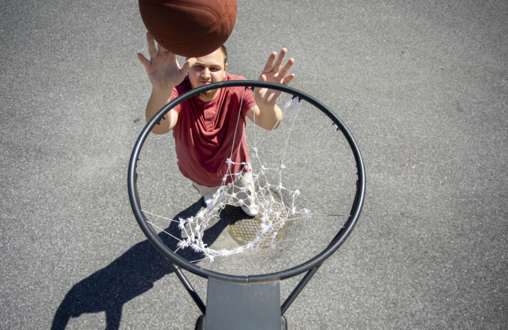 Man in red shirt, arms outstretched above head shooting an orange basketball toward a metal hoop while playing in his driveway.