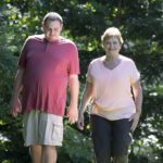 Man in red shirt and tan cargo shorts walks alongside woman in pink top and black pants in a wooded yard