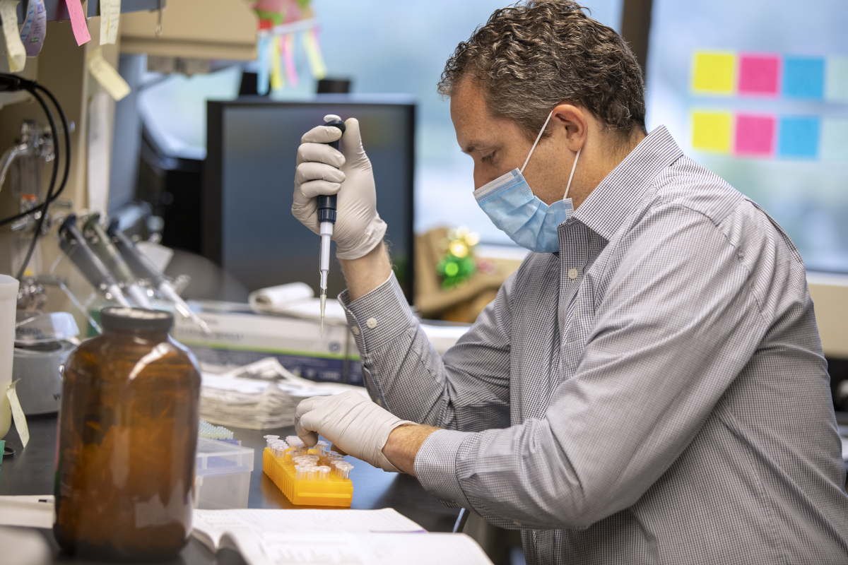 Virologist Nick Buchkovich, seated at a lab bench, uses pipetting equipment. Additional lab equipment and various books and papers are also on the bench.