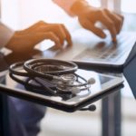 A physician types on a laptop. A stethoscope and iPad are next to the laptop.