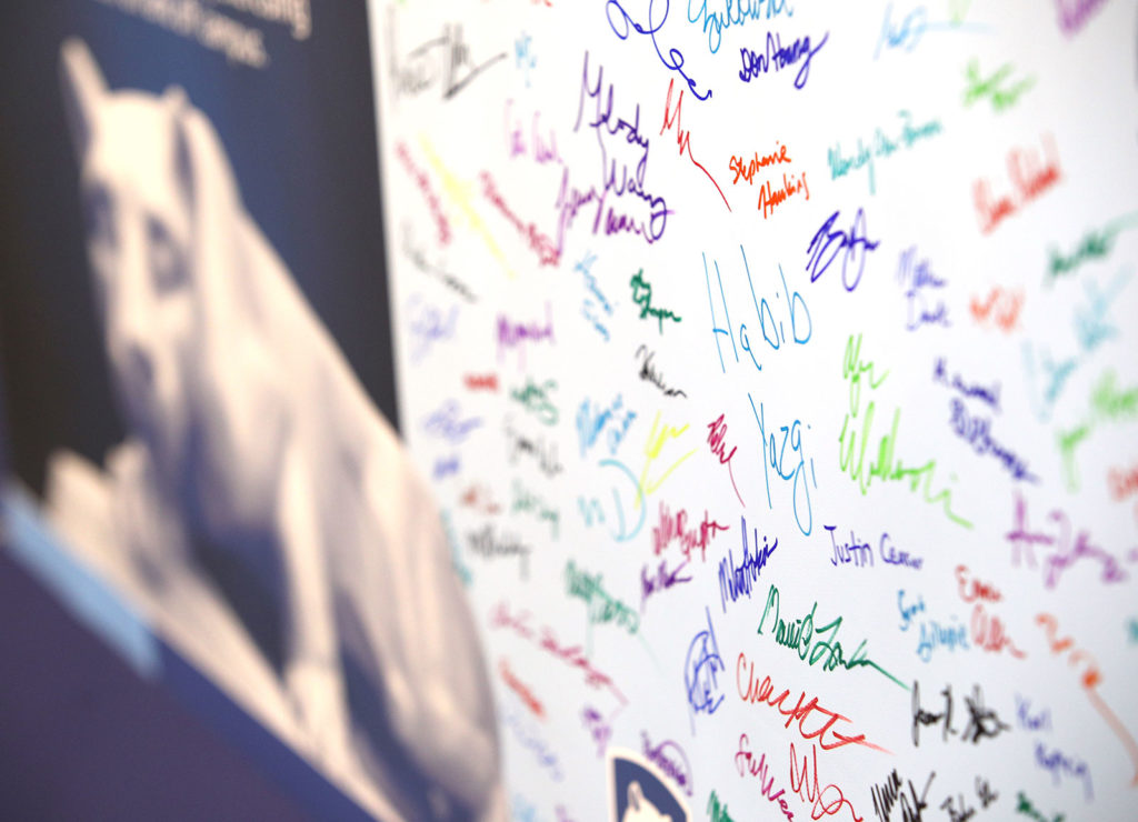 A close-up image shows signatures in many colors of marker on a large poster. The Penn State Nittany Lion is visible out of focus to the left.