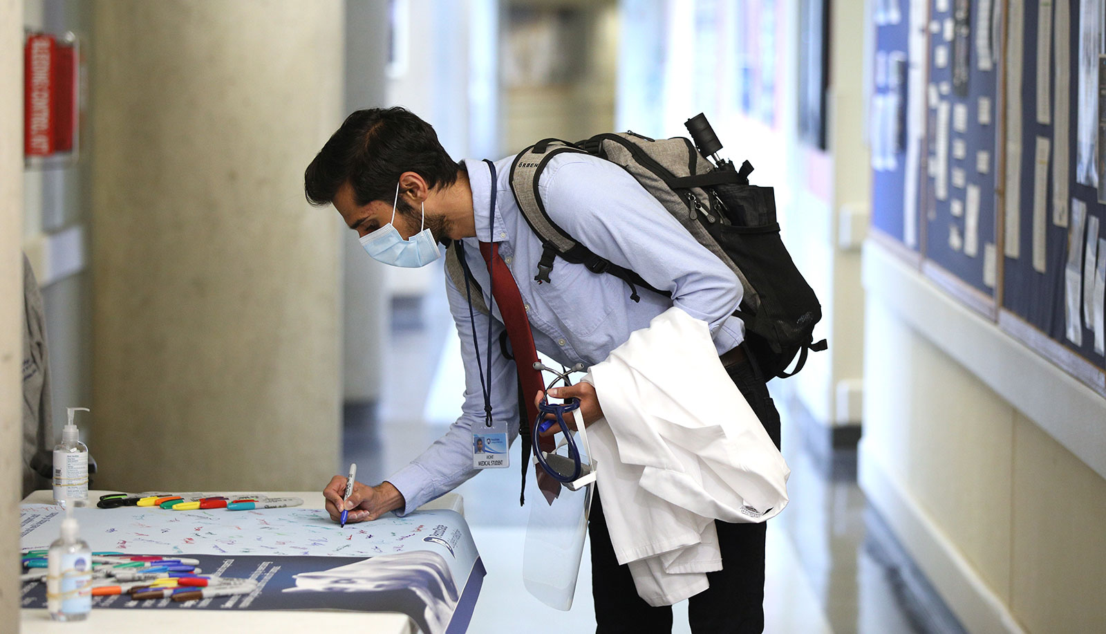 A man wearing a face mask and carrying a white medical student coat uses a marker to sign a large banner laying on a table.