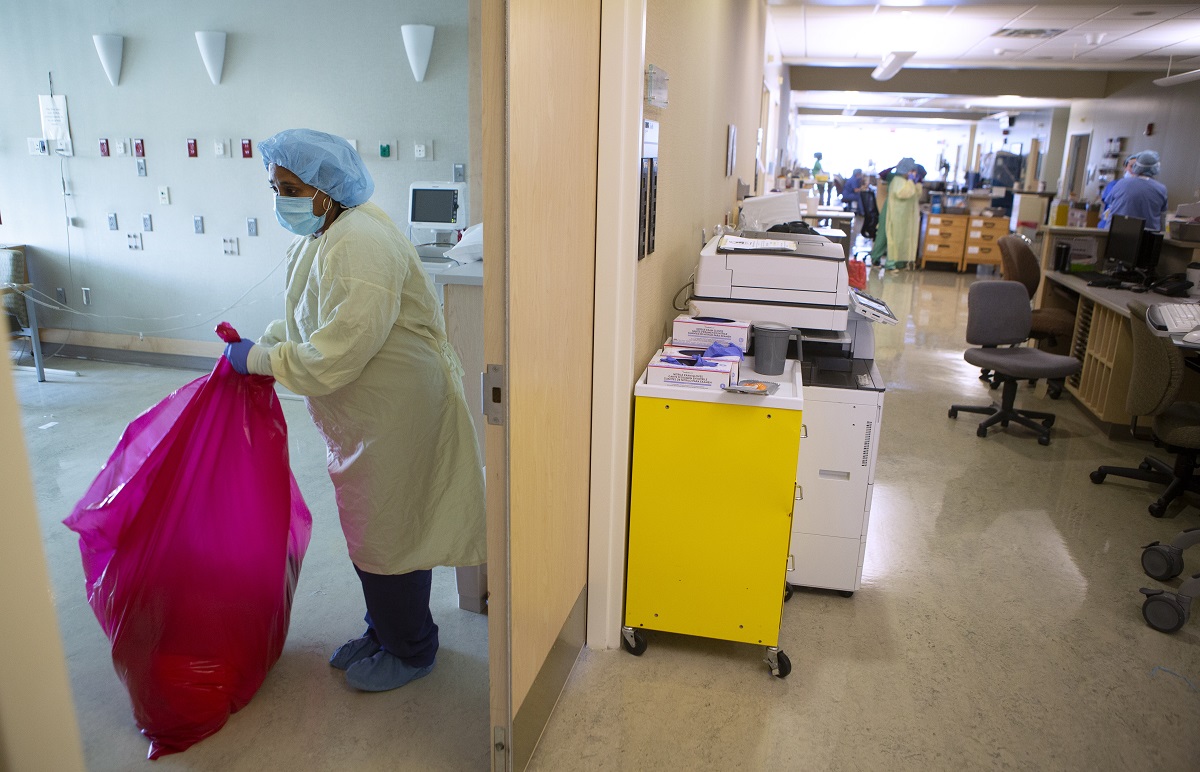 Woman standing in yellow gown, blue hair cover, blue facemask, blue gloves and blue shoe covers holds a red bag while removing red-bag waste from a patient room.
