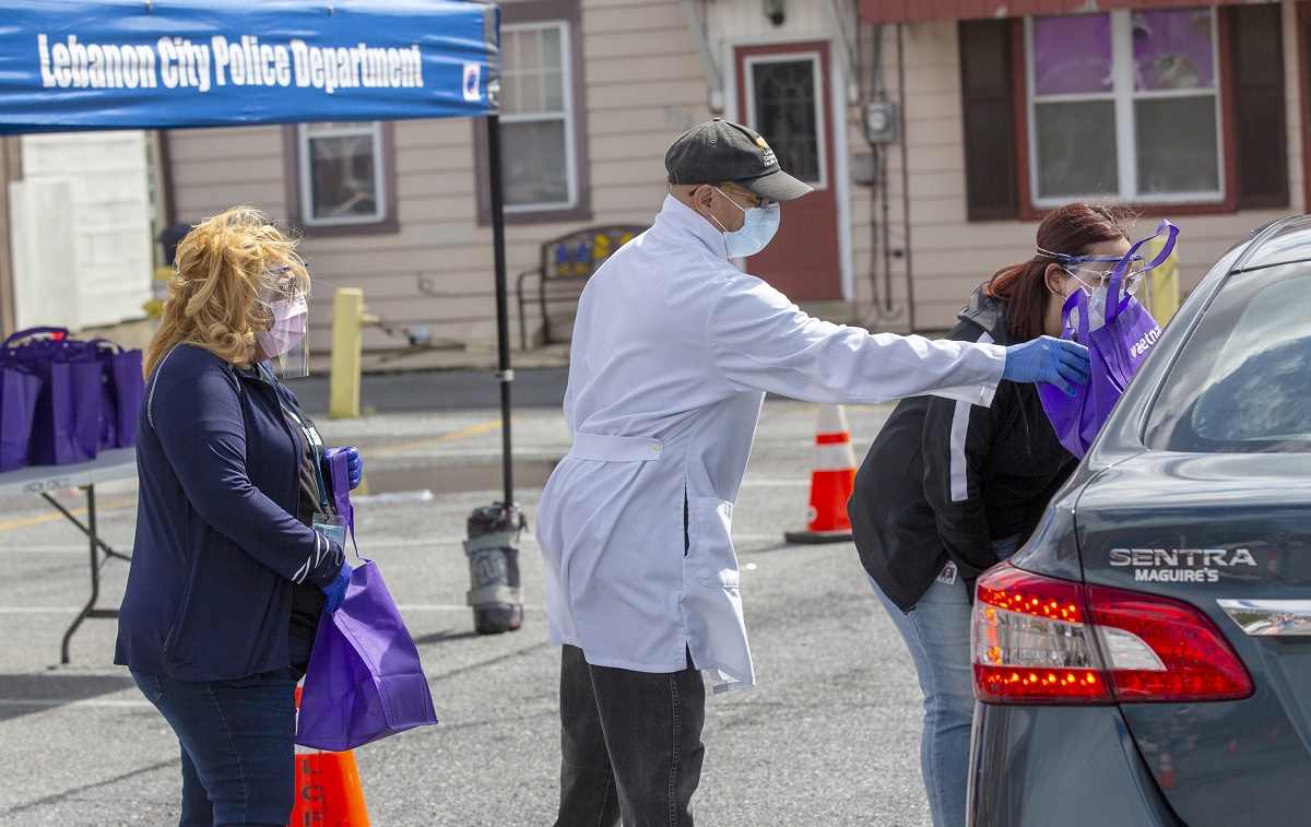 Two women and one man hand a bag to a driver in a car. All three are wearing face masks. The man is wearing a white coat and baseball cap. Behind them is a pop-up tent with the words “Lebanon City Police Department” on it.