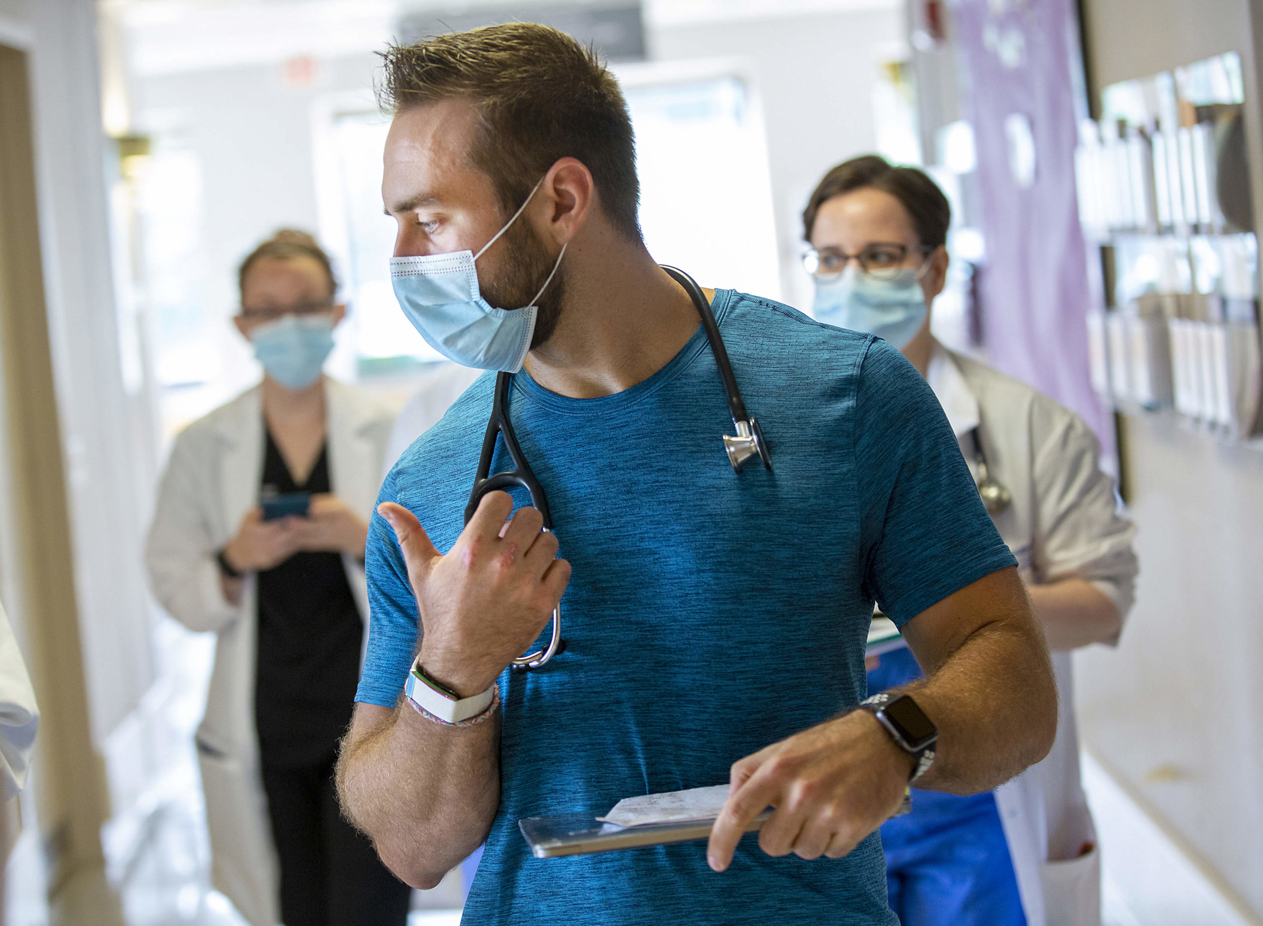Dr. Alexander Hajduczok, a third-year internal medicine resident at Penn State Health Milton S. Hershey Medical Center, sports a wearable device manufactured by WHOOP on his right wrist. Two other physicians are behind him, walking down a hallway.