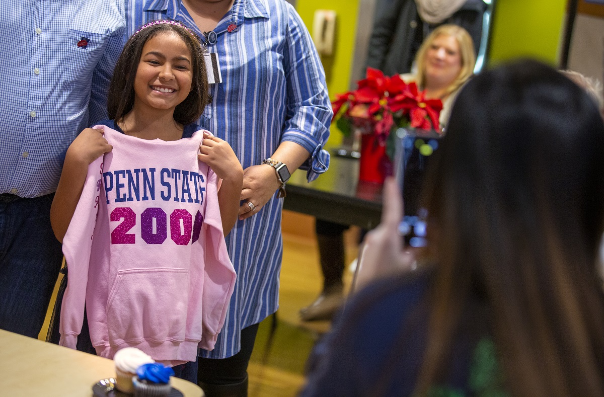 Ruth Vega smiles as she holds a sweatshirt up in front of herself that says Penn State 2000th. Her parents stand behind her. A poinsettia plant sits on a table in the background, with people sitting around it.