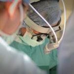 El Dr. Zakiyah Kadry, con una red para el cabello, un sombrero quirúrgico, una máscara facial y una bata quirúrgica, mira hacia abajo en la mesa de operaciones. Otro miembro del personal está cerca de la cámara, fuera de foco.