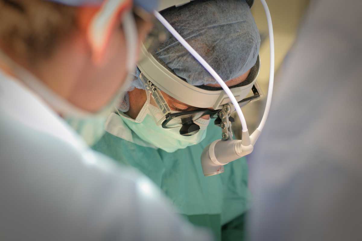 El Dr. Zakiyah Kadry, con una red para el cabello, un sombrero quirúrgico, una máscara facial y una bata quirúrgica, mira hacia abajo en la mesa de operaciones. Otro miembro del personal está cerca de la cámara, fuera de foco.