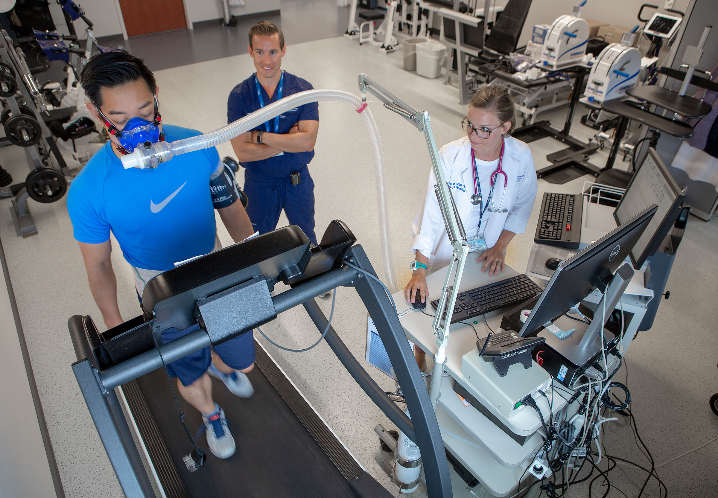 A patient walks on a treadmill wearing a face mask in the lab of Dr. Jonathan Stine at Penn State Health Milton S. Hershey Medical Center. Dr. Stine and exercise physiologist Brei Hummer are watching the patient.