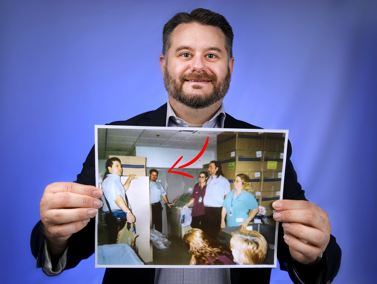 Kristofer Miller, who has a beard and moustache and is smiling, holds up a picture of a group of people. A red arrow drawn on the picture shows him among six co-workers, who are standing by stacks of large cardboard boxes and laundry bins.