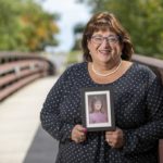 Carolyn Powell, who wears glasses and is smiling, smiles and holds a picture of herself taken in her early days at Hershey Medical Center. She is wearing a polka-dotted blouse and a string of pearls and stands on a bridge with trees in the background.