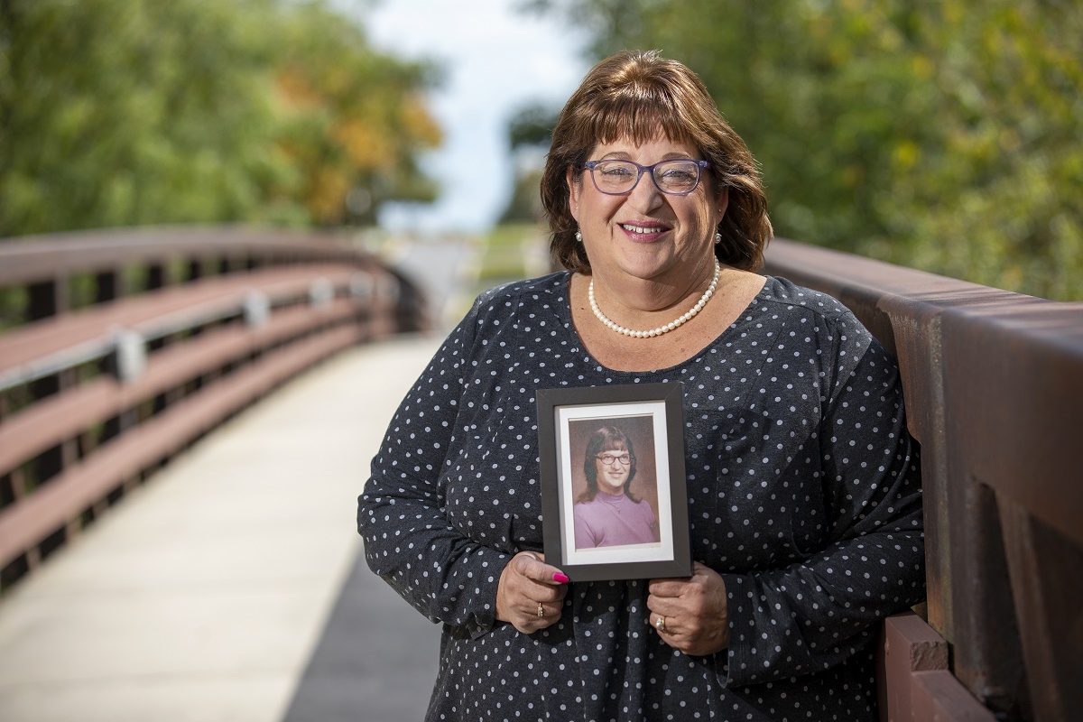 Carolyn Powell, who wears glasses and is smiling, smiles and holds a picture of herself taken in her early days at Hershey Medical Center. She is wearing a polka-dotted blouse and a string of pearls and stands on a bridge with trees in the background.