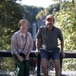 Glenna and J. David Brensinger lean against the side of a bridge. In the background are trees and, at a distance, a large waterfall.