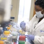 Jasmine Geathers is working in a lab at Penn State College of Medicine. She is wearing a white coat with the Penn State Hershey logo on it, a face mask and gloves. She is holding an instrument in a container of test tubes. A table full of bottles and other lab equipment is in front of her.