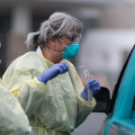 Two women in personal protective equipment, including gowns, masks, goggles, and gloves, stand alongside a vehicle, speaking to the driver, who’s not visible. One worker holds a swab.