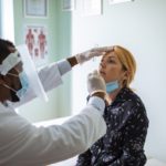 A health care worker uses a swab to get a sample from a woman’s nostril. The health care worker wears personal protective equipment.