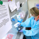 A woman dressed in personal protective equipment is seated at a lab bench over which there is an exhaust hood. A "precautions" sign is in the foreground.