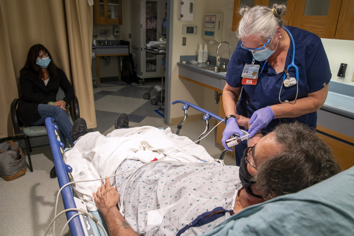 A female nurse administers medication at the bedside of a male patient in an Emergency Department room at Hershey Medical Center. A woman is seated in a chair nearby, looking on.