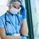 A health care worker wearing scrubs, rubber gloves, a hair net and a surgical mask raises her hand to her forehead and wears an exasperated look.