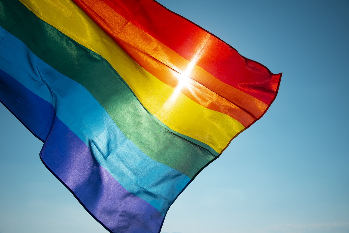 A flag with rainbow stripes waves in front of the sun.