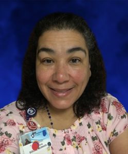 Gloria Rosado, a social worker at Penn State Health St. Joseph — Downtown Campus, smiles at the camera in a professional headshot. She is wearing a flowered dress, a lanyard with a Penn State Health ID badge and necklace. She has shoulder-length, curly hair.