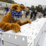The "Nittany Lion," wearing a facemask, uses a marker to sign a metal beam. Several people walk around in the background.