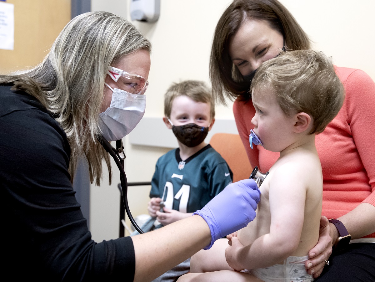Dr. Melissa Tribuzio, wearing glasses and a mask, holds a stethoscope to 2-year-old Matthew Fitzpatrick’s chest as he sits on his mother’s lap. He wears a diaper and has a pacifier in his mouth. Samantha Fitzpatrick, who is wearing a sweater and a mask, places her hand behind his back. His little brother, Ben, is in the background and wears a mask.