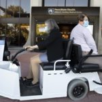 Two women drive in a golf cart in front of the Hershey Medical Center entrance. The woman on the left is driving and wearing a skirt, jacket and mask. The woman on the right is wearing a long-sleeved top, pants and a mask.