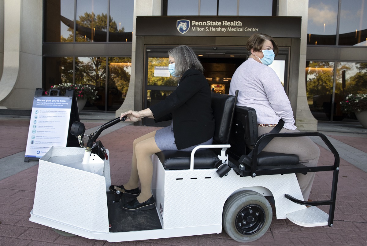 Two women drive in a golf cart in front of the Hershey Medical Center entrance. The woman on the left is driving and wearing a skirt, jacket and mask. The woman on the right is wearing a long-sleeved top, pants and a mask.