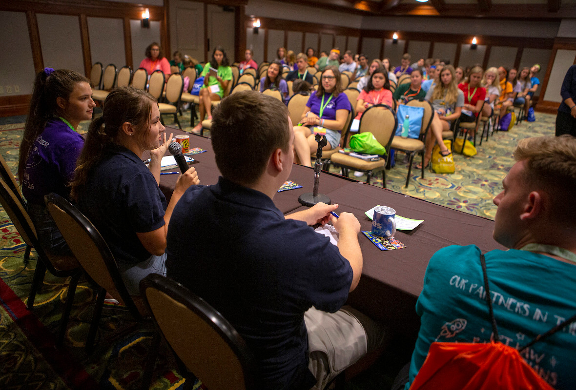 Students sit at a table and speak to a crowded room