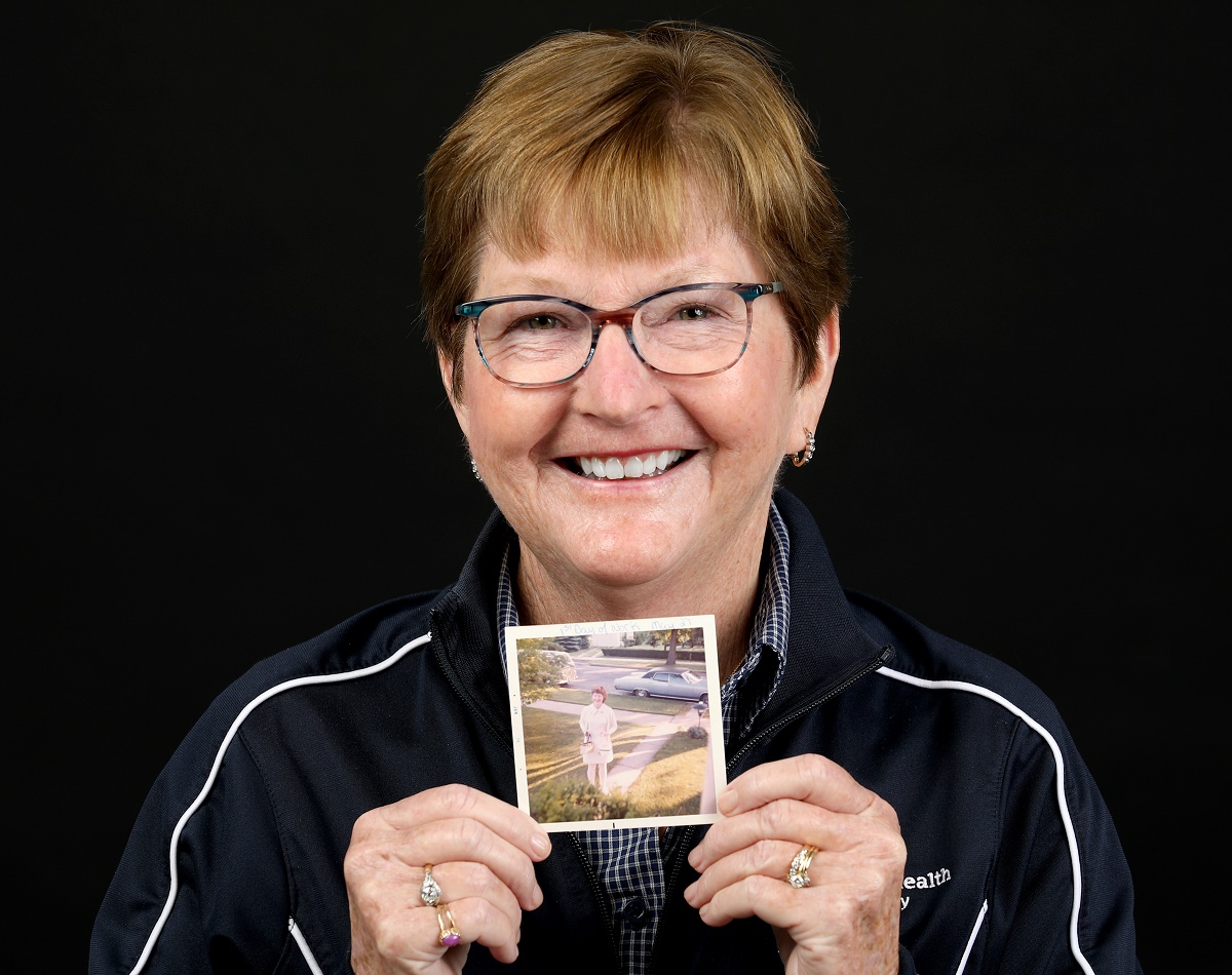 Janice Mills, who has short hair and glasses, smiles and holds a vintage picture of herself in a nurse’s uniform.