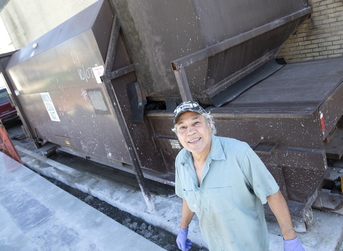 Alex Astier, wearing a short-sleeve shirt, ball cap and rubber glove, stands by a dumpster and smiles.