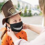 La joven vestida para Halloween con sombrero de copa marrón y camisa de calabaza consigue su máscara facial de tela negra de cara sonriente ajustada por su madre antes de salir para “trick or treat.”