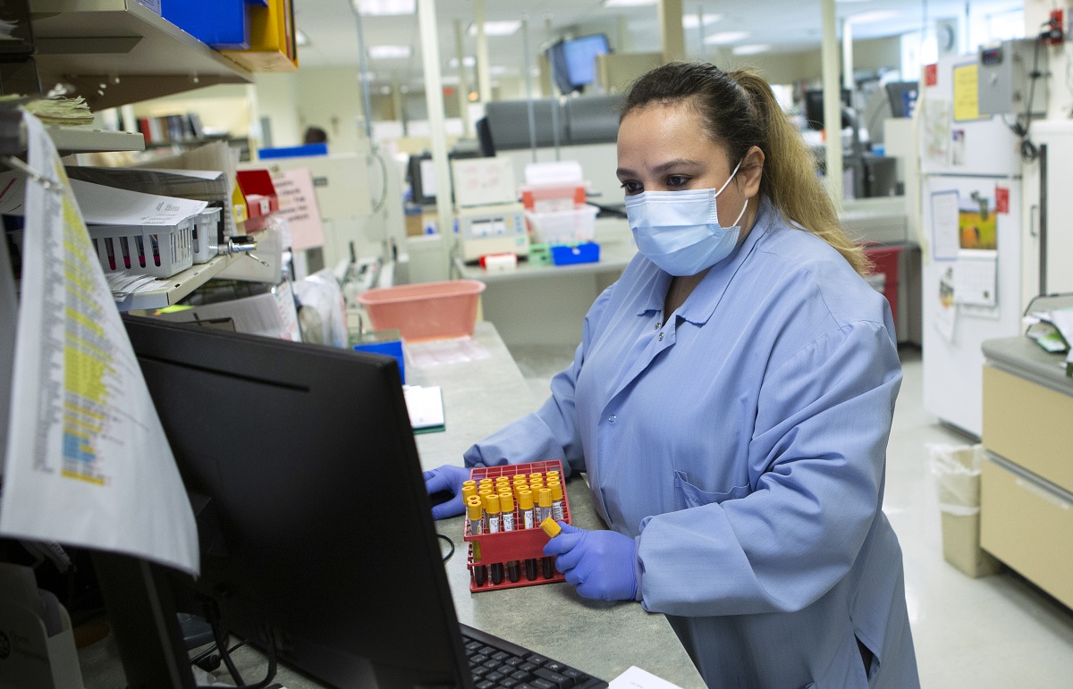 Osiris Martinez Urquilla looks at a computer screen as she stands in the medical laboratory at Penn State Health St. Joseph Medical Center. She is holding a test tube and is wearing scrubs, gloves and face mask and has long hair in a ponytail.
