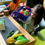 Savannah Hoover holds a plastic basket of toy groceries during a field trip to Whitaker Center for the Science and Arts in Harrisburg. She stands beside a toy check-out counter, filled with plastic toy food and milk cartons.