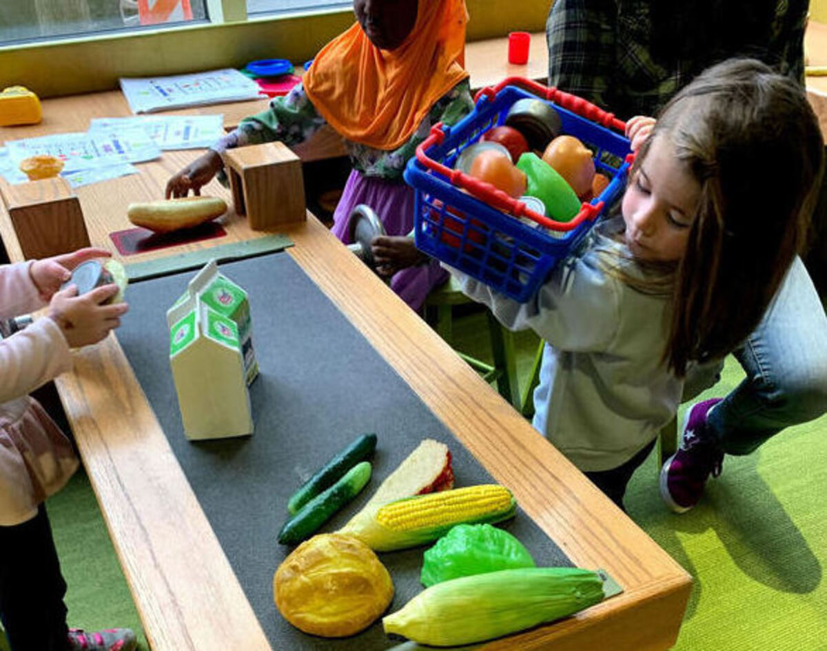 Savannah Hoover holds a plastic basket of toy groceries during a field trip to Whitaker Center for the Science and Arts in Harrisburg. She stands beside a toy check-out counter, filled with plastic toy food and milk cartons.