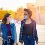 Two women, each wearing face masks, walk along a street side by side, looking at each other.