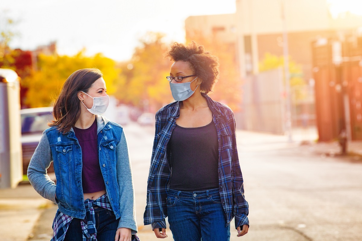 Two women, each wearing face masks, walk along a street side by side, looking at each other.